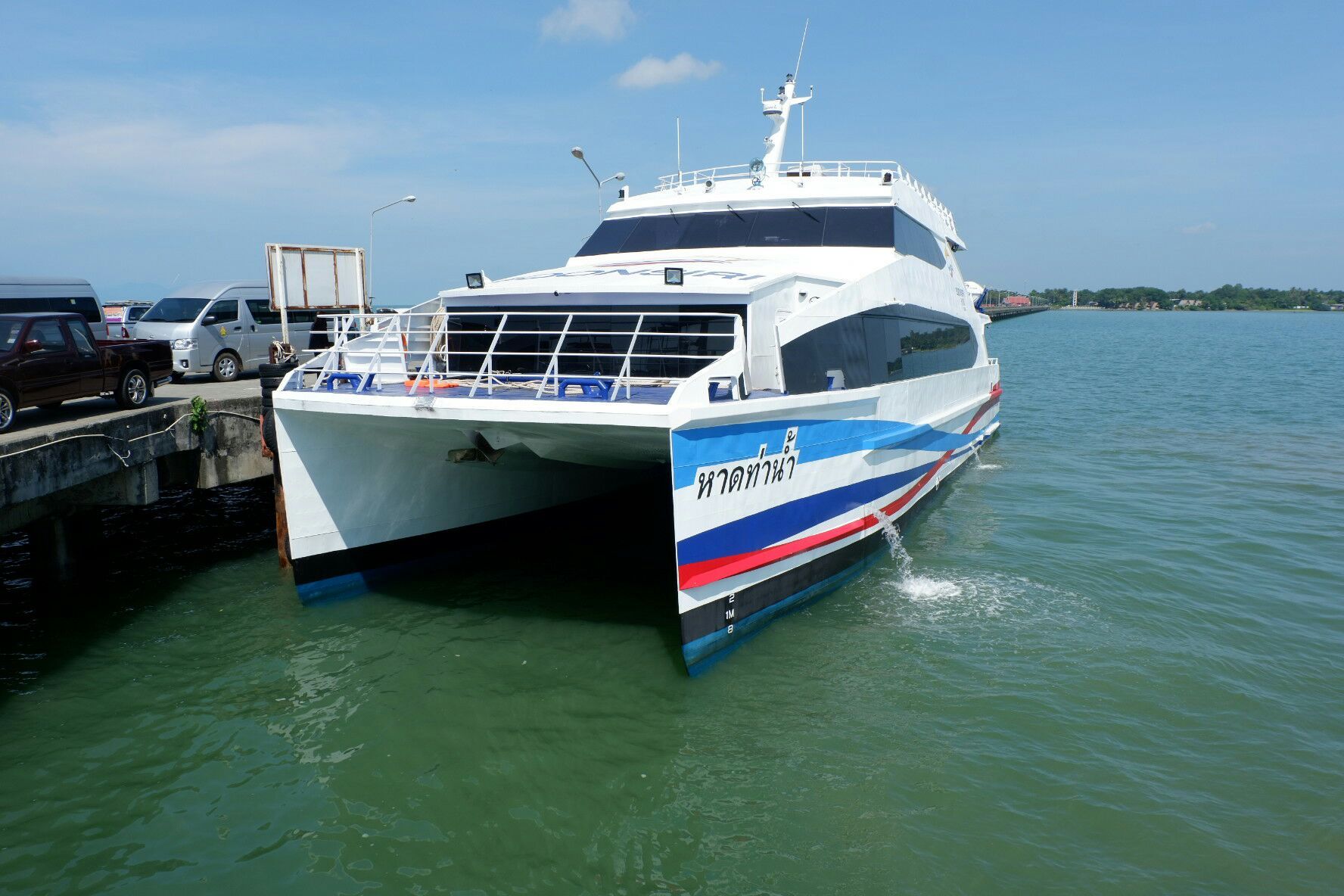 Catamarã rápido branco com letras tailandesas no casco atracado num cais de betão sob céu azul