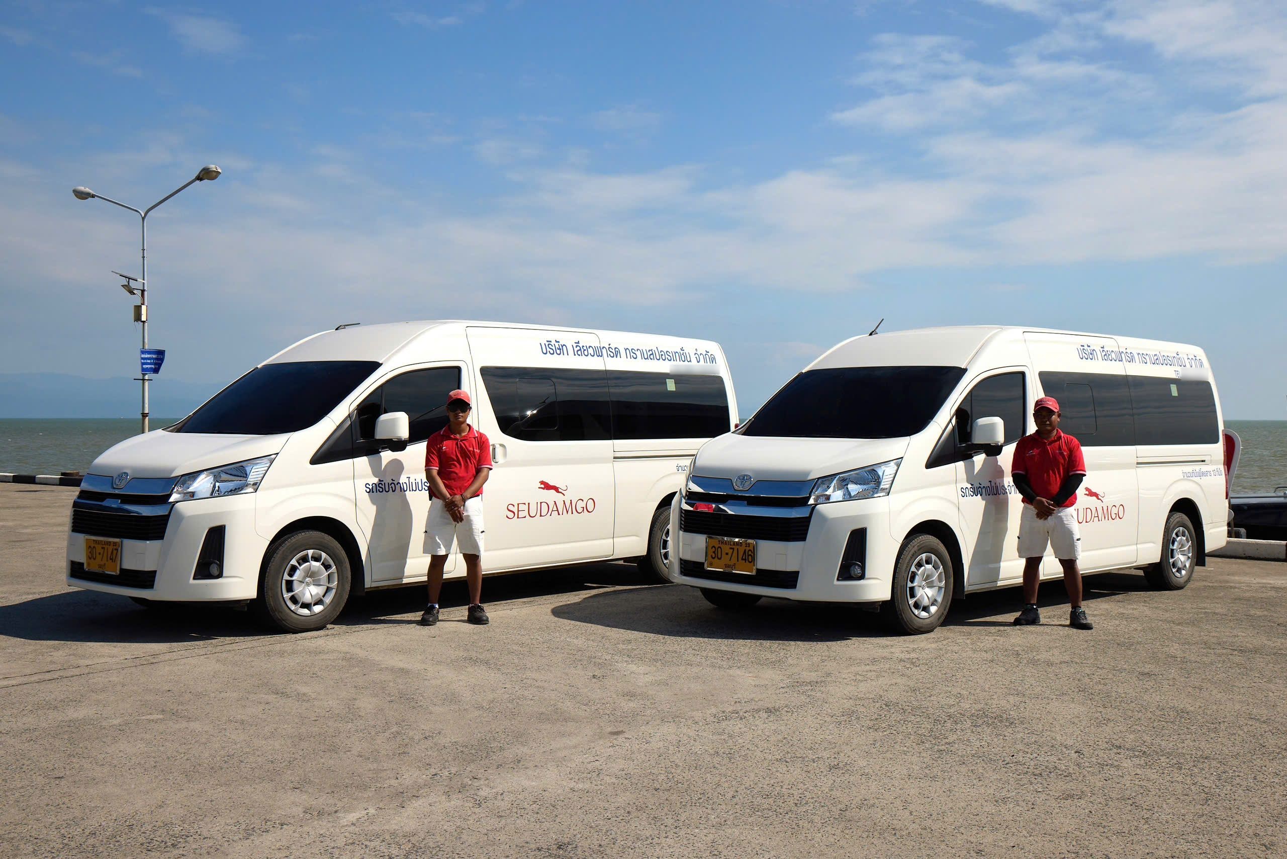 Duas carrinhas Toyota brancas com marca vermelha num cais de betão junto ao mar, motoristas de camisa vermelha ao lado