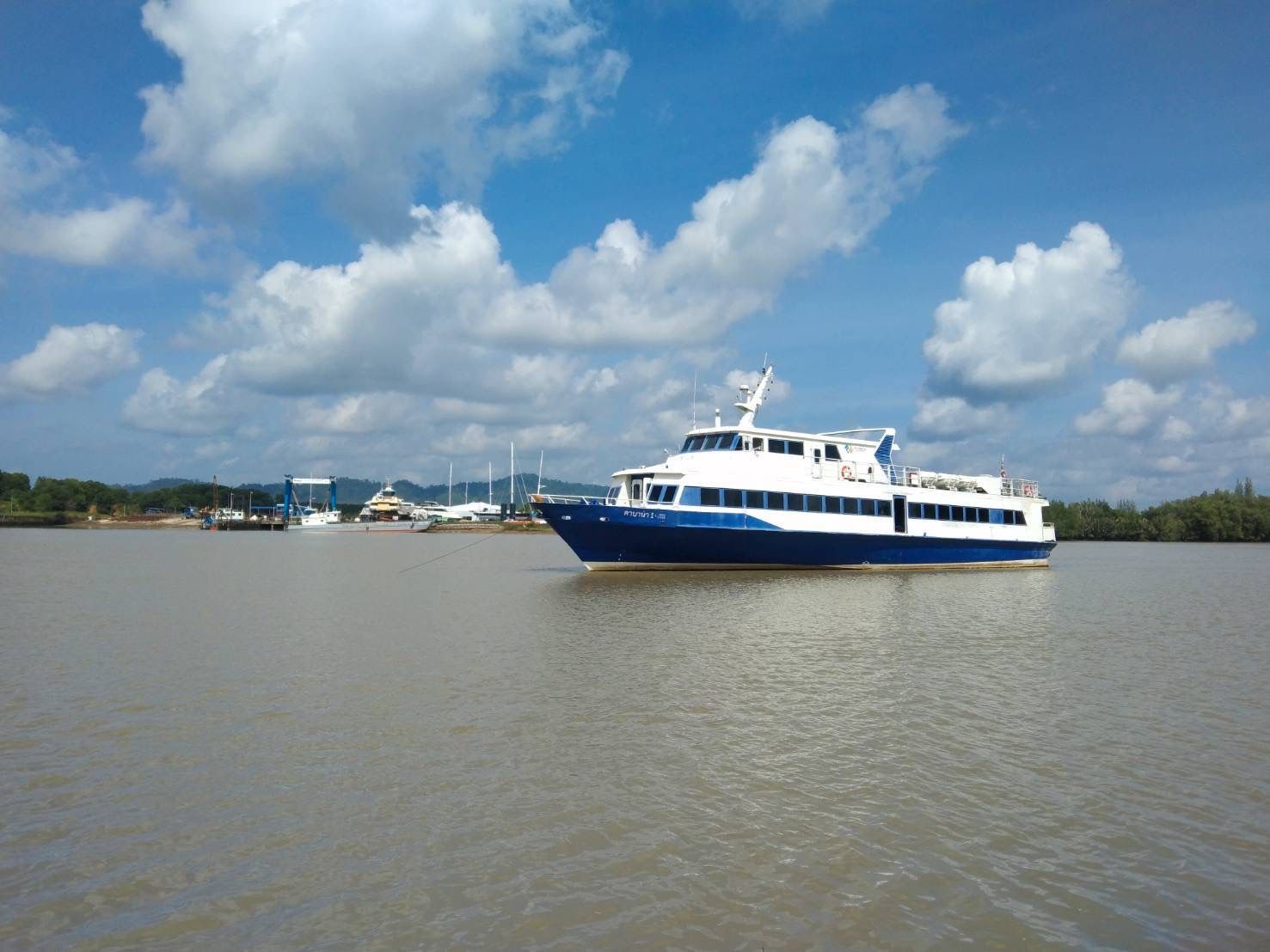 Ferry de passageiros azul e branco fundeado num rio de maré castanho junto a mangais, com pequena marina atrás