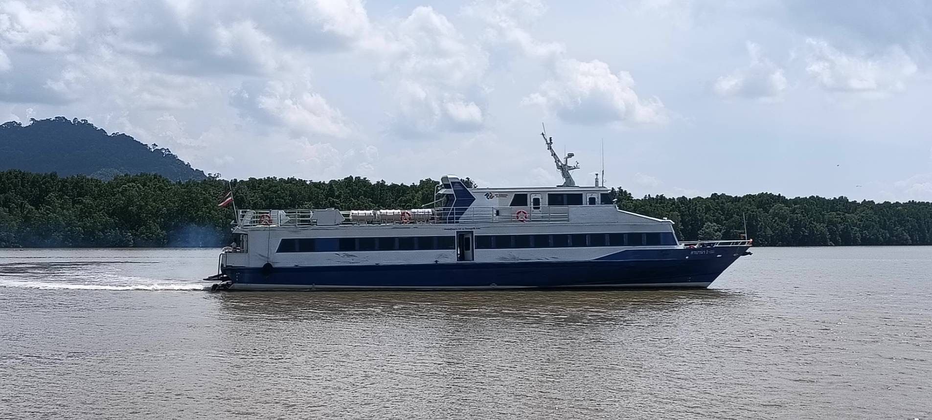 Ferry de passageiros azul e branco a navegar num estuário castanho junto a mangais densos e colinas, céu nublado