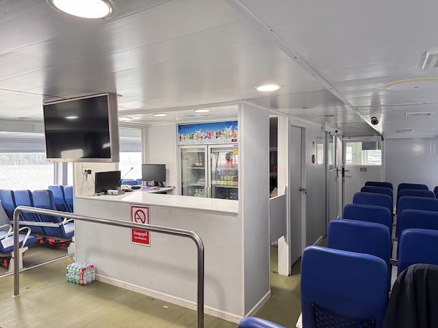 Interior da cabina do ferry com bancos azuis, balcão de snacks, arca de bebidas, ecrã de TV e sinal de proibido fumar