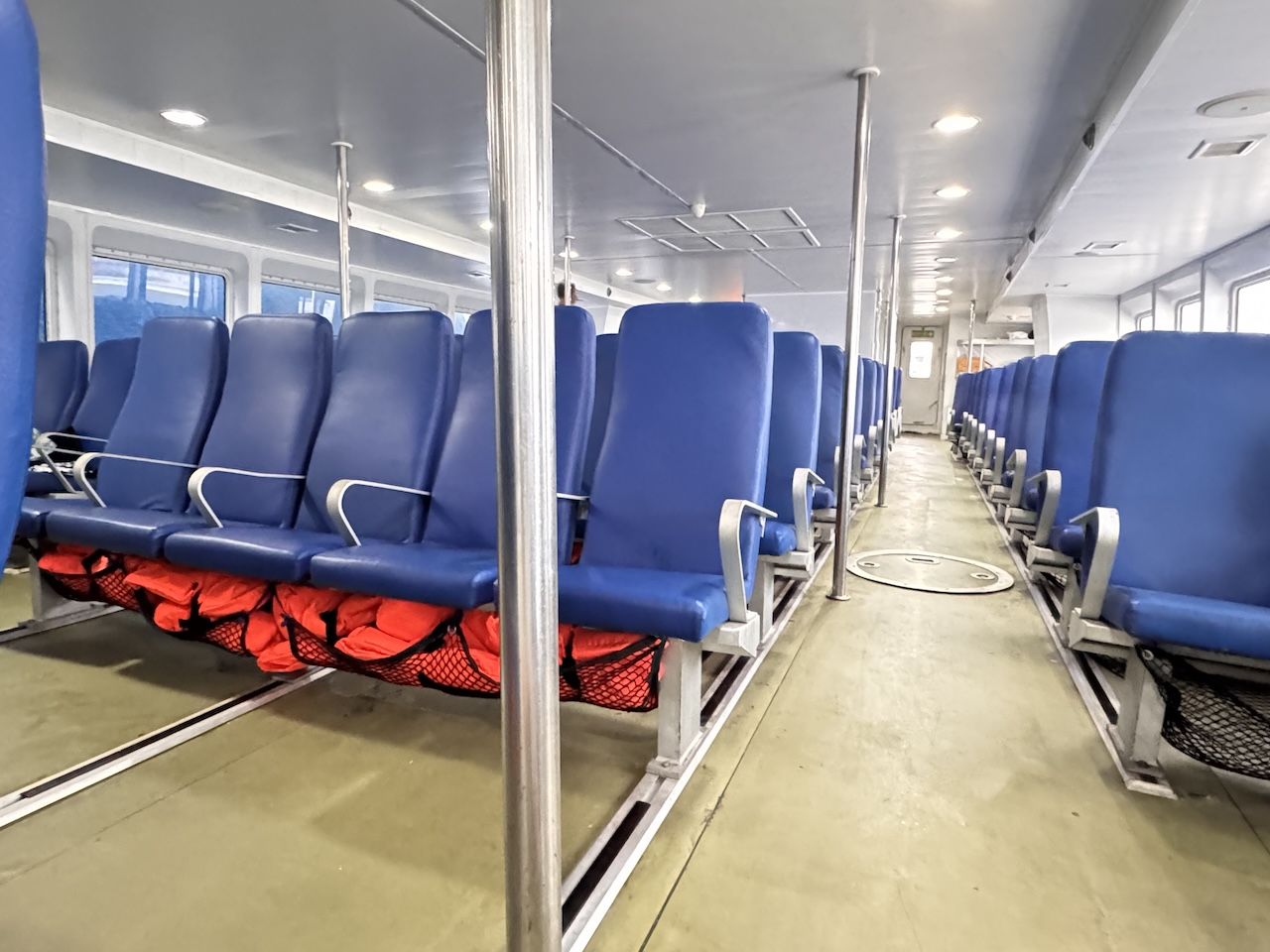 Cabina de passageiros vazia de ferry com filas de bancos azuis, coletes salva-vidas laranja em redes e corredor central
