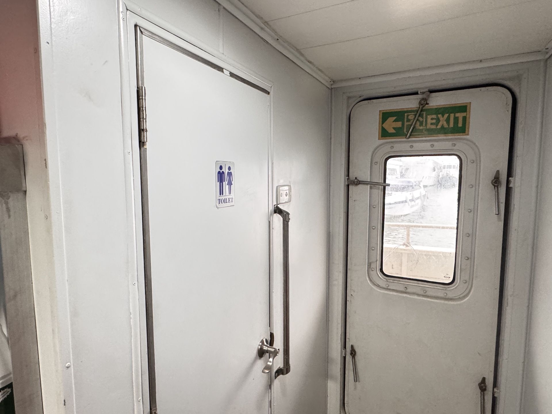 Corredor interior de ferry com porta de casa de banho branca e porta estanque de saída com sinal verde EXIT