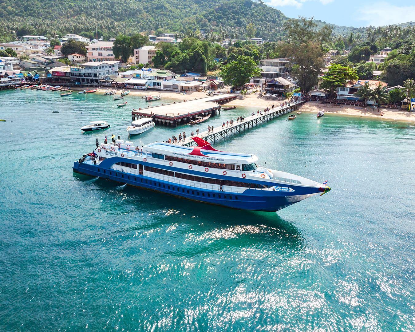 Vista aérea de um ferry azul e branco a aproximar-se de um cais com turistas, baía de ilha tropical