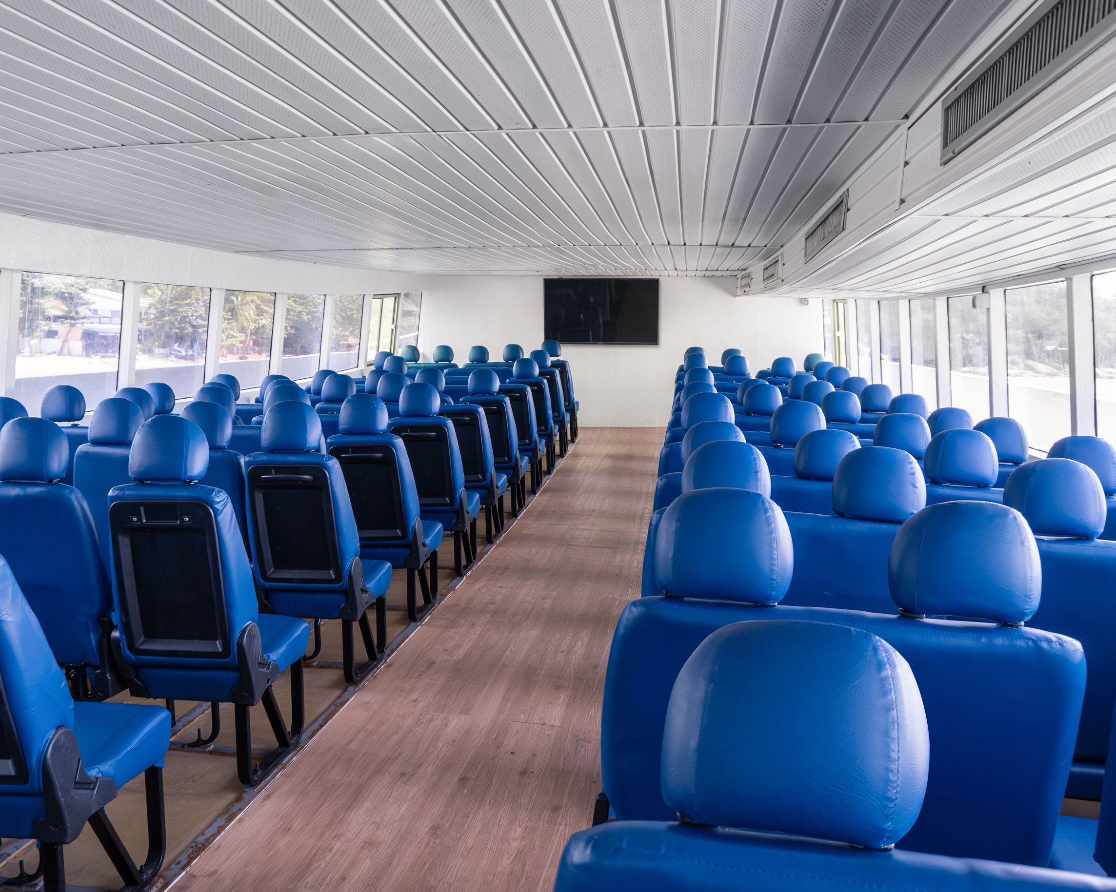 Cabina de passageiros vazia de ferry com filas de bancos azuis, corredor central em madeira e janelas amplas