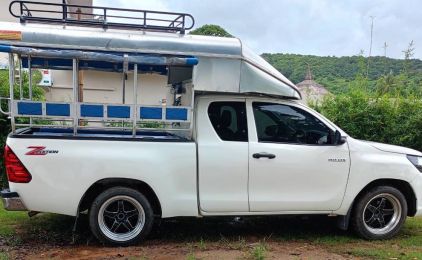 Pickup branca convertida em songthaew com bancos corridos, toldo e barras de tejadilho, na relva junto a colinas verdes