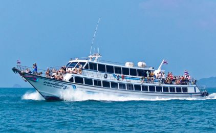 Ferry de passageiros branco e cheio, com bandeiras tailandesas, navegando em mar turquesa perto de uma ilha arborizada
