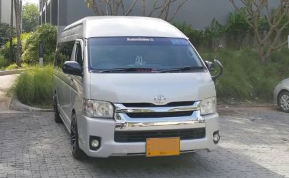 รถตู้หลังคาสูง Toyota Hiace สีเงิน จอดบนทางรถเข้าปูพื้นข้างสนามหญ้าและอาคารสมัยใหม่