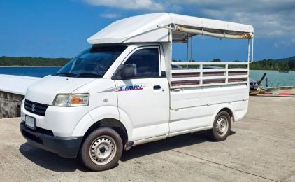 Pickup Suzuki Carry branca com banco de passageiros coberto, estacionada num cais de betão junto ao mar tropical calmo