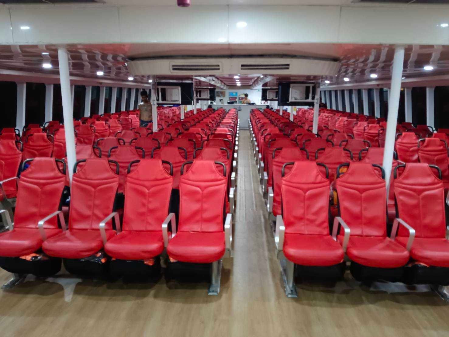 Cabina de passageiros de ferry vazia com filas de bancos vermelhos, corredor central e chão imitação madeira