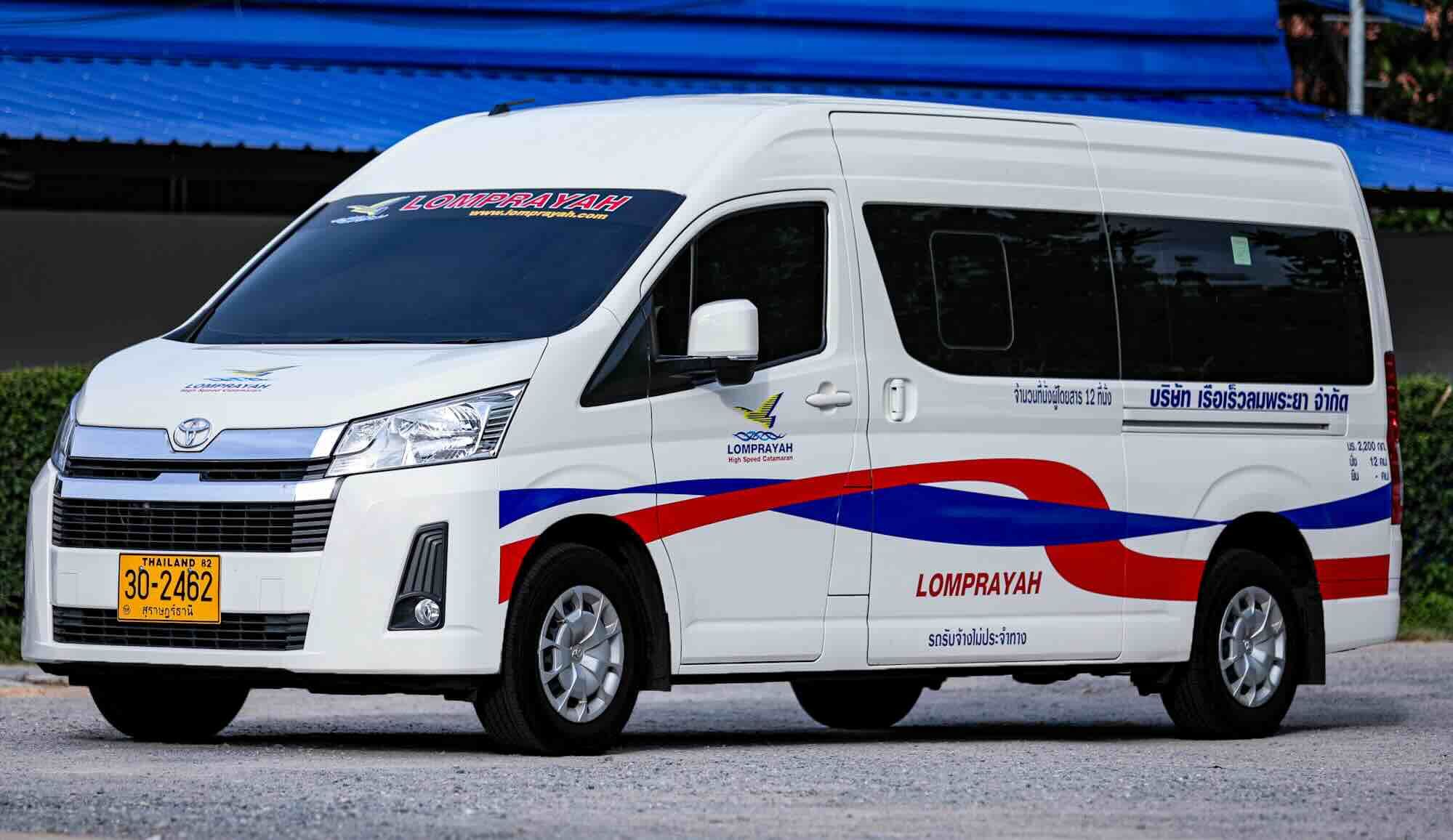 White Toyota Hiace minivan with Lomprayah livery and Thai plates parked on gravel by a blue roof