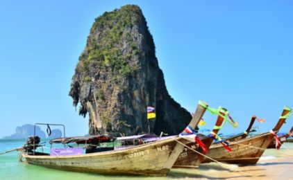 Barcos longtail tailandeses tradicionais com fitas coloridas atracados numa praia sob um enorme rochedo calcário