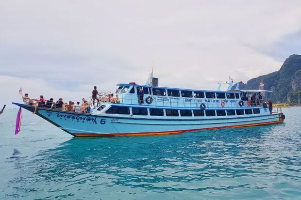 Ferry de passageiros tailandês azul e branco com pessoas a apanhar sol no convés da proa a cruzar mar turquesa junto a falésias calcárias