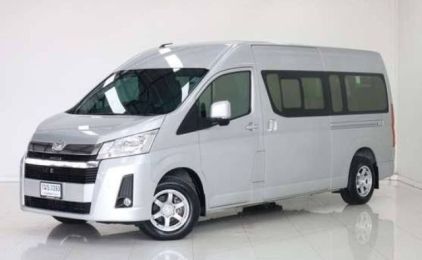Carrinha de passageiros Toyota Hiace prateada estacionada num showroom interior iluminado, vista frontal de três quartos