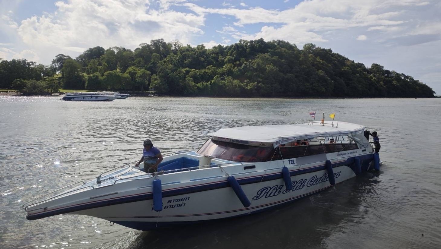 Lancha branca com letras tailandesas em água pouco profunda junto a ilha arborizada, céu parcialmente nublado