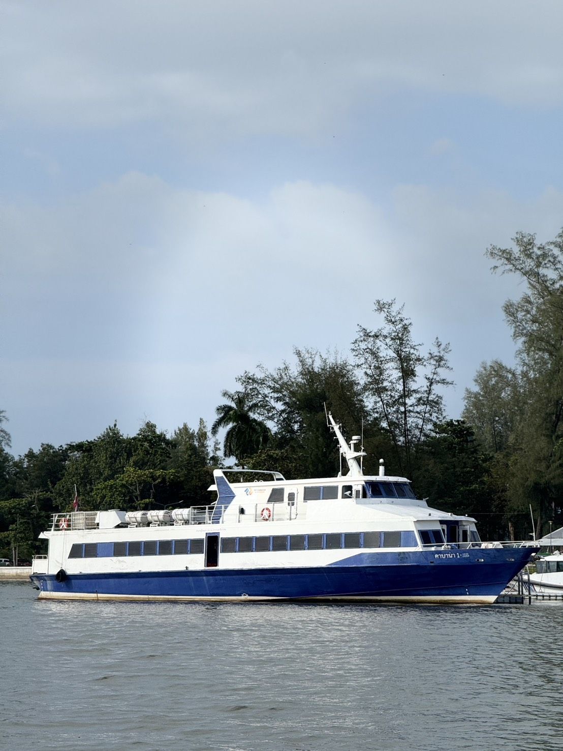 Ferry de passageiros azul e branco atracado num cais, com letras tailandesas na proa e pinheiros atrás