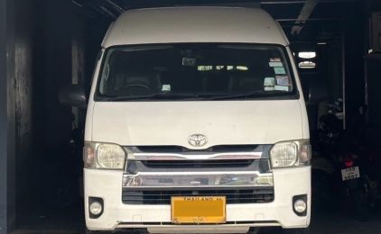 รถตู้โดยสาร Toyota Hiace สีขาวจอดหันหน้าเข้าหากล้องในโรงรถแสงสลัว พร้อมป้ายทะเบียนไทยสีเหลือง