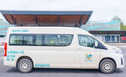 Furgoneta branca de teto alto com letras tailandesas e logótipo de agência de viagens estacionada sob o alpendre de um terminal