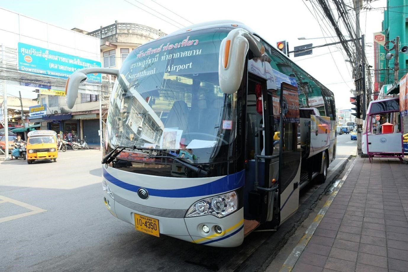 Flughafen-Shuttlebus von Suratthani auf einer thailändischen Straße, dahinter Tuk-Tuks und Ladenschilder
