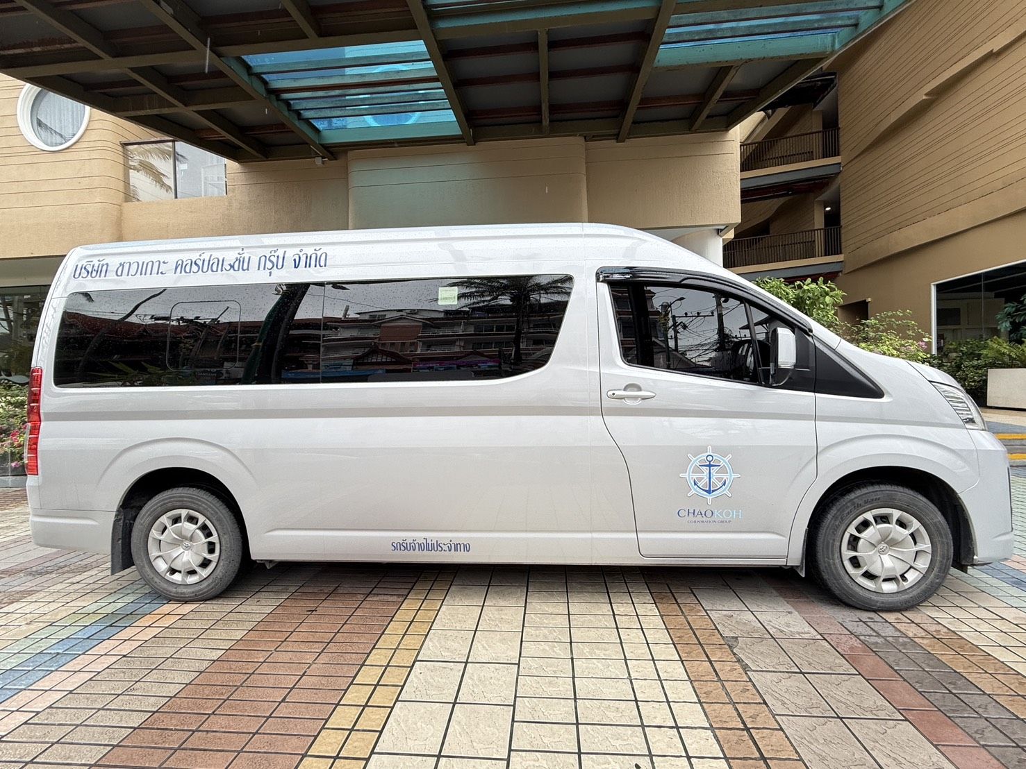 Carrinha Toyota Hiace branca com letras tailandesas e logótipo de âncora do Chao Koh Corporation Group sob toldo de hotel
