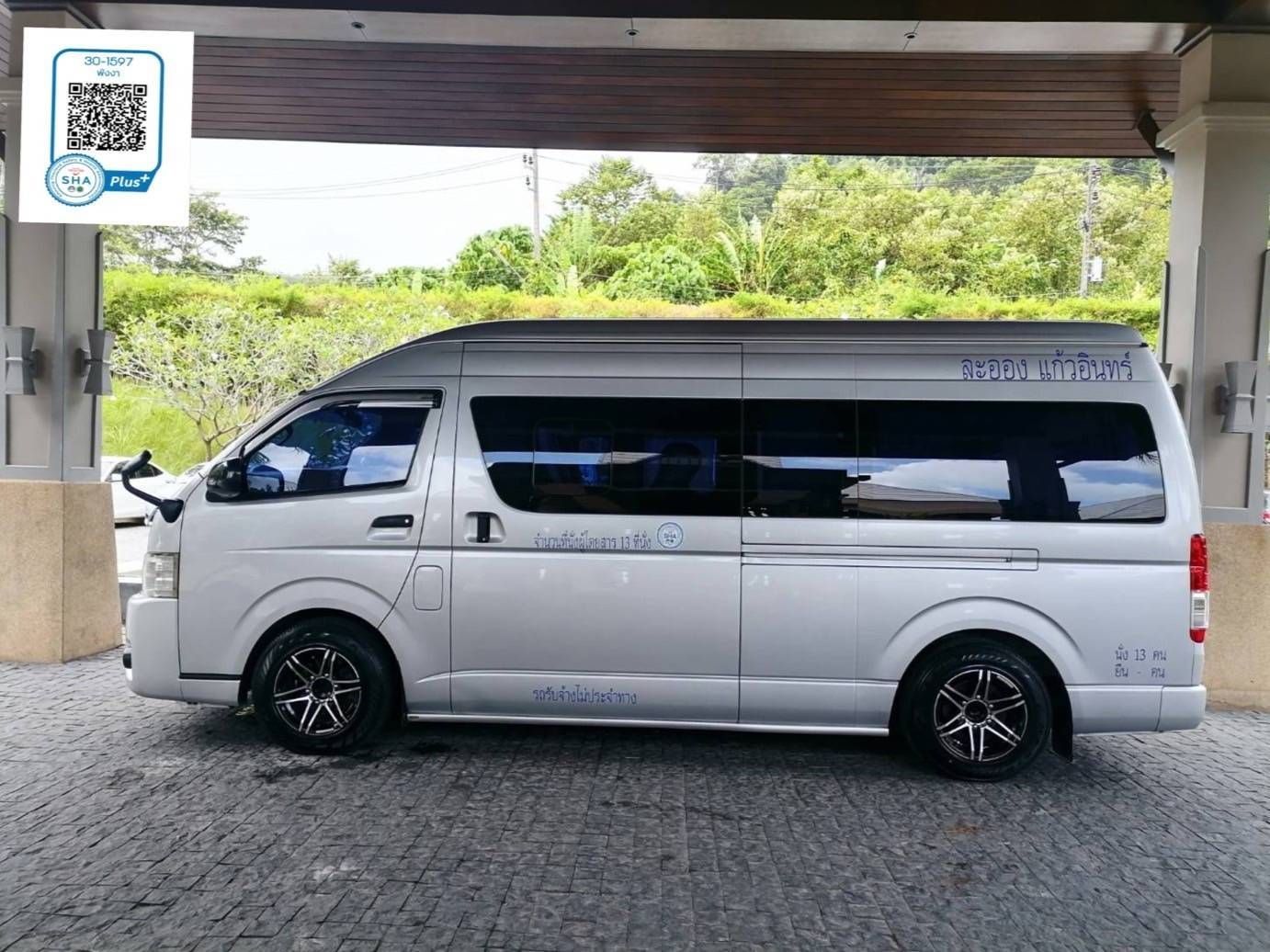 Carrinha Toyota Hiace prateada de 13 lugares com letras tailandesas sob alpendre de hotel em Phang Nga