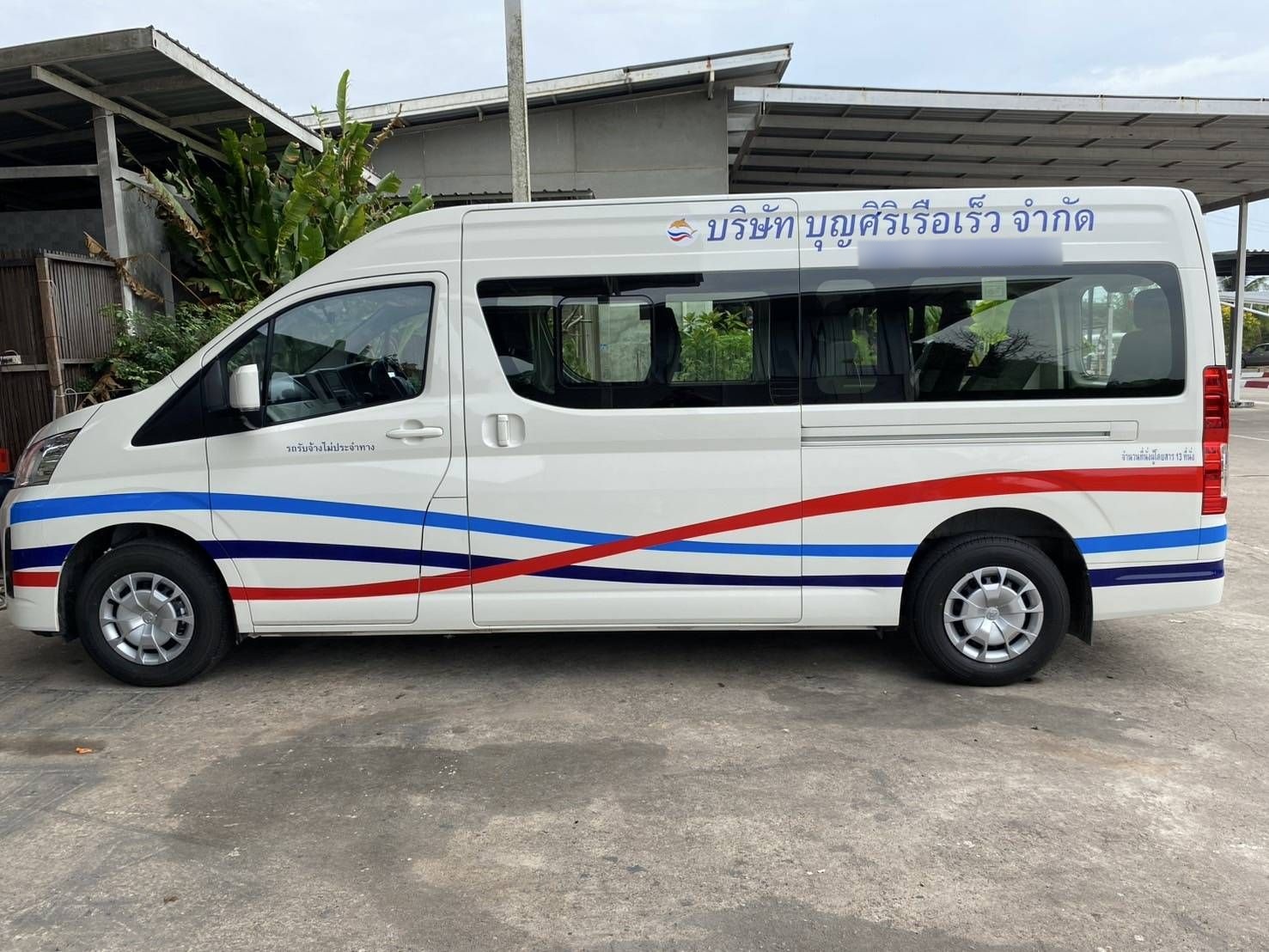 Minibus branco com faixas curvas vermelhas e azuis e inscrições em tailandês, estacionado em cimento à saída de um alpendre