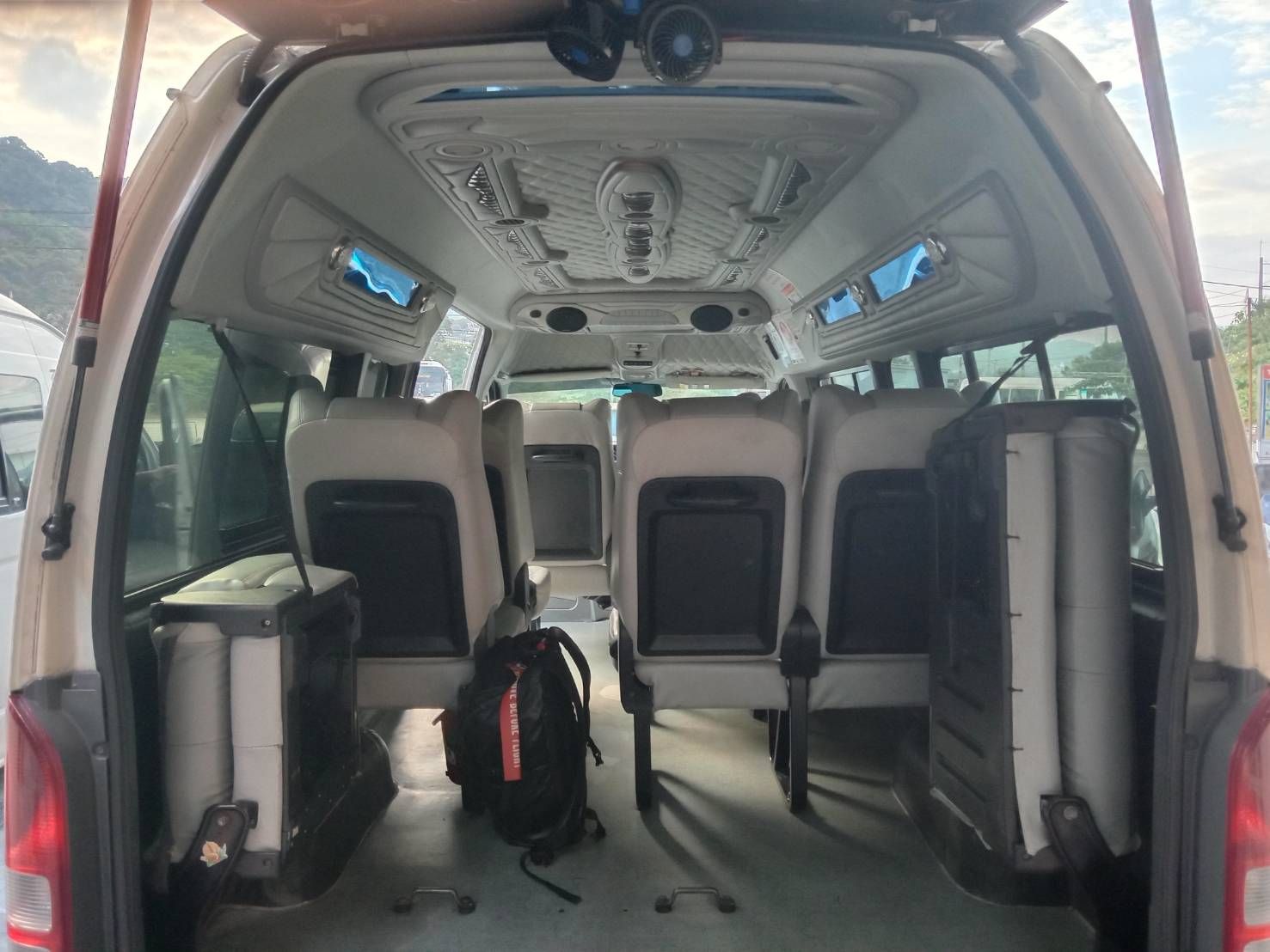 Interior de uma minivan visto pelas portas traseiras abertas, bancos creme, ventoinhas no teto e uma mochila preta no chão