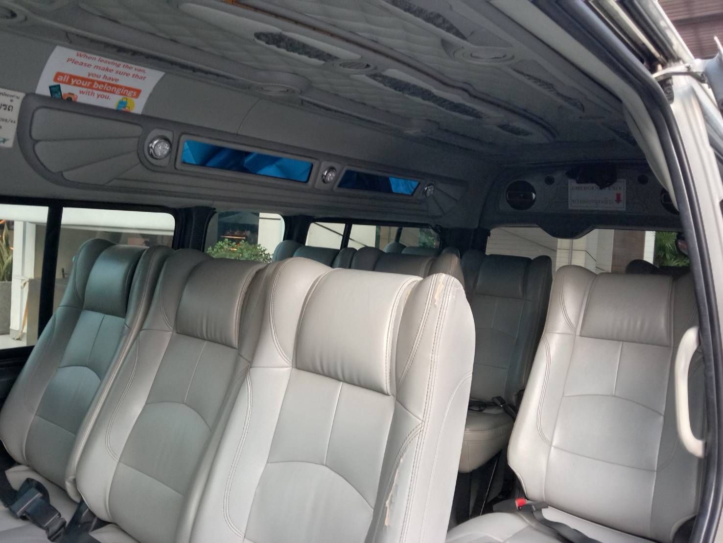 Interior de uma minivan de passageiros com bancos de pele cinzenta, saídas de ar no teto e um autocolante informativo em tailandês e inglês