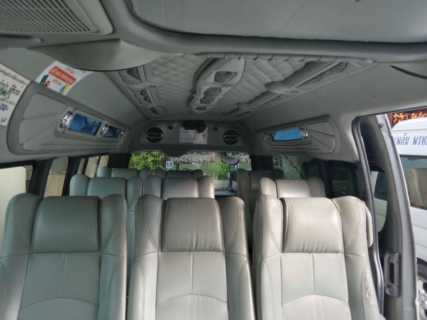 Interior de uma minivan de passageiros com filas de bancos de pele cinzenta, porta lateral aberta e sinalética tailandesa visível no exterior