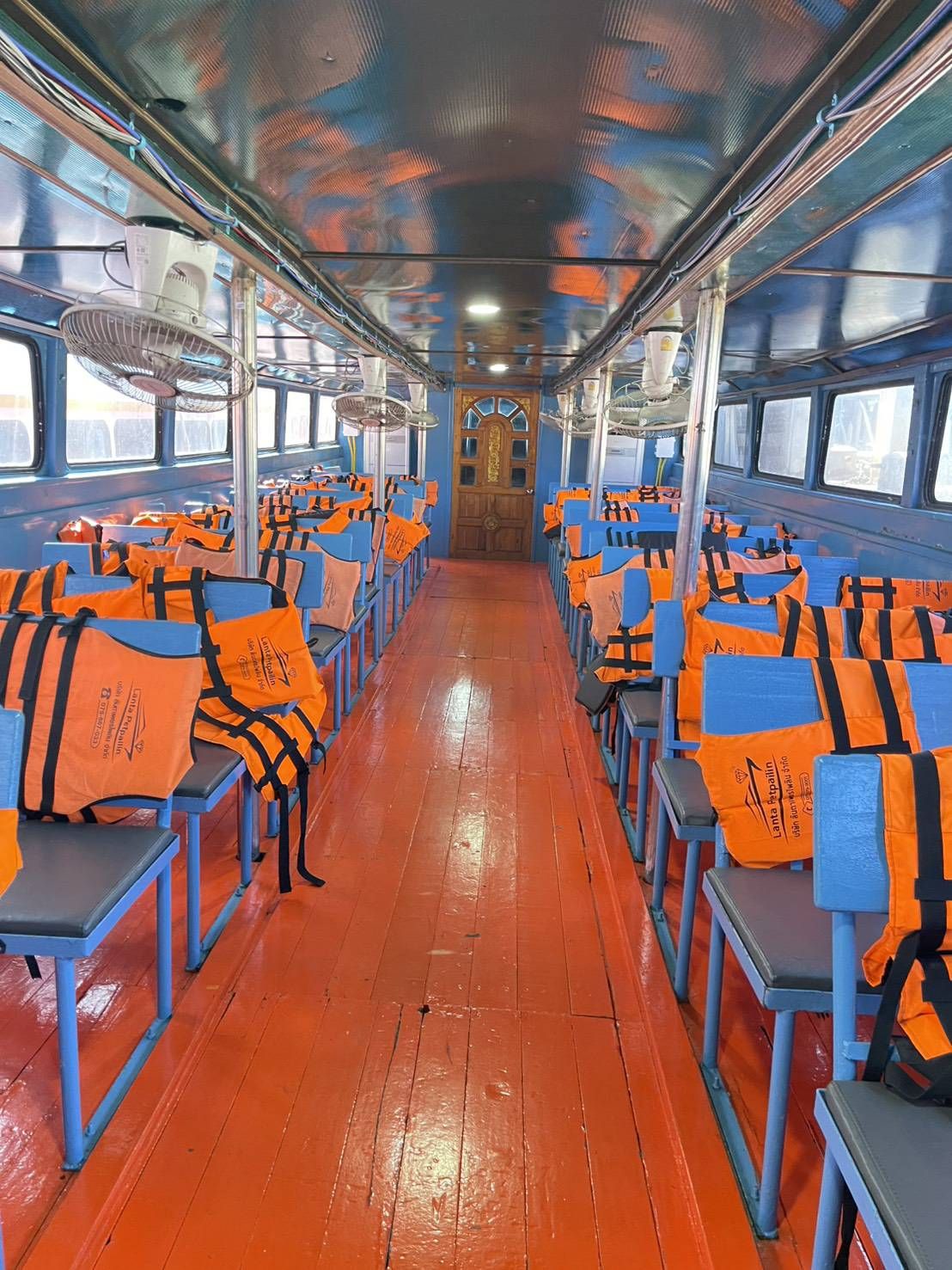 Cabina de passageiros vazia de ferry com coletes salva-vidas laranja em bancos azuis, piso de madeira laranja e ventoinhas de teto