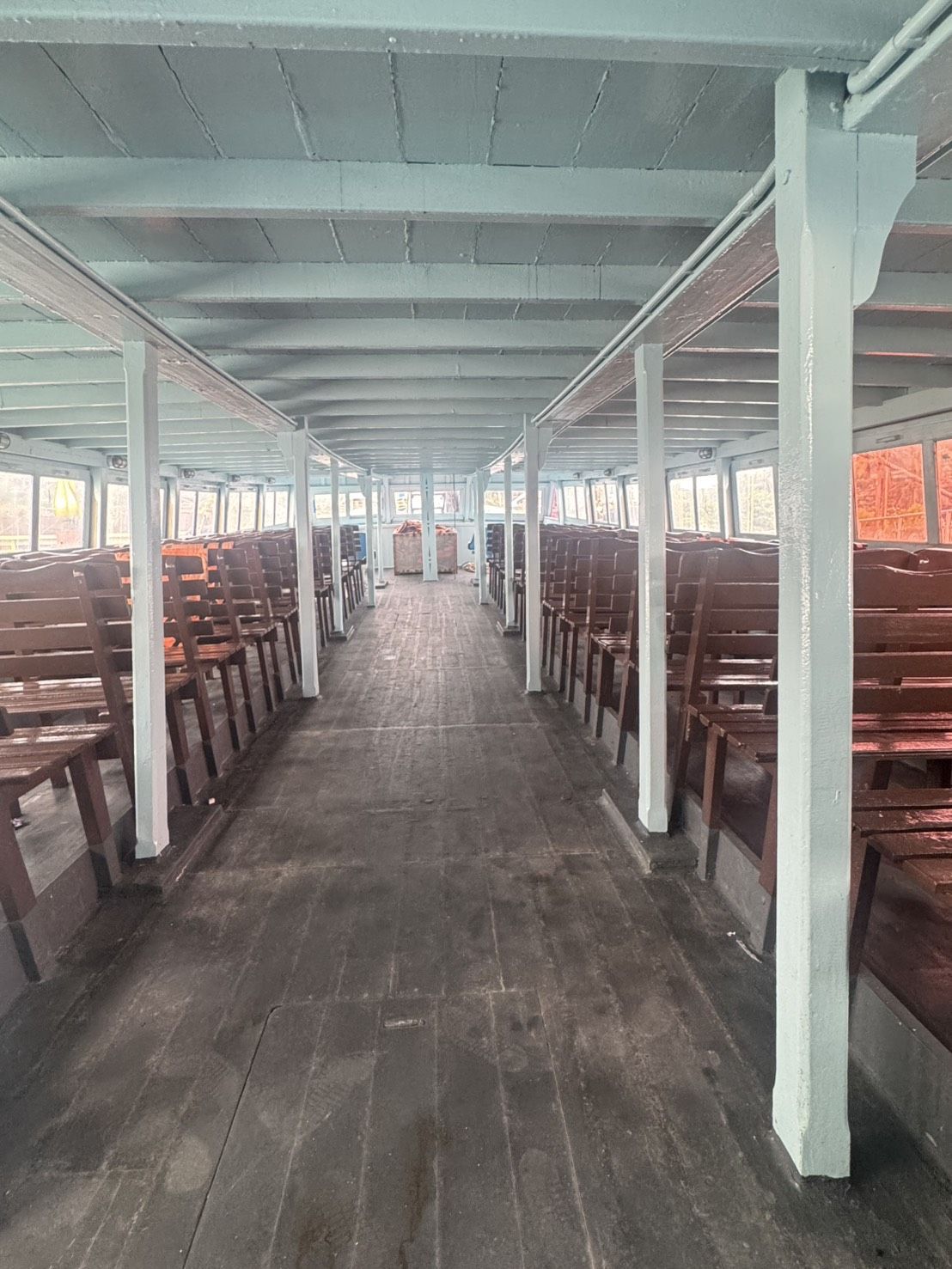 Deque de passageiros vazio de um ferry de madeira com filas de bancos marrons, colunas azul-claras e janelas abertas