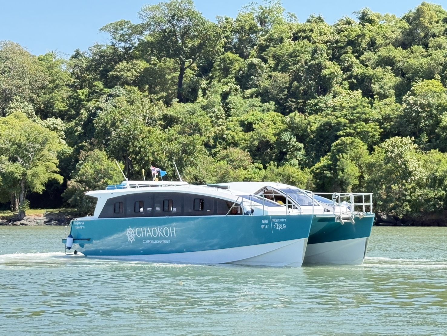 Lancha catamarã Chaokoh azul-petróleo e branca a navegar em águas verdes calmas junto à costa de uma ilha arborizada