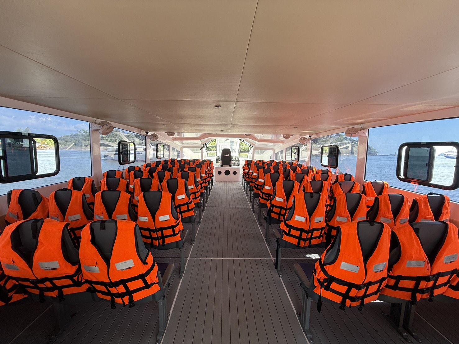 Cabina de passageiros vazia de uma lancha ferry com filas de bancos cobertos por coletes salva-vidas laranja e vista para o mar