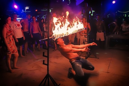 Artista de fogo a cuspir chamas num evento noturno com espectadores sob luzes de palco coloridas