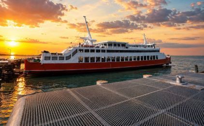 Ferry de passageiros vermelho e branco atracado numa rampa metálica ao pôr do sol, com nuvens laranja sobre o mar calmo