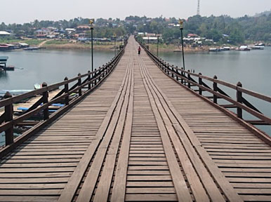 Longa ponte pedonal de madeira a atravessar um rio calmo em direção a uma aldeia na encosta, na Tailândia